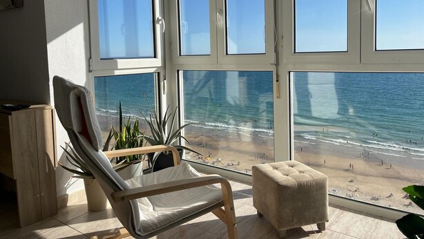 Ein Zimmer mit großem Fenster mit Blick auf einen Strand, ein gepolsteter Sessel und ein kleiner Hocker.