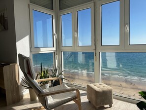 A room with a large window overlooking a beach, a cushioned chair, and a small ottoman.