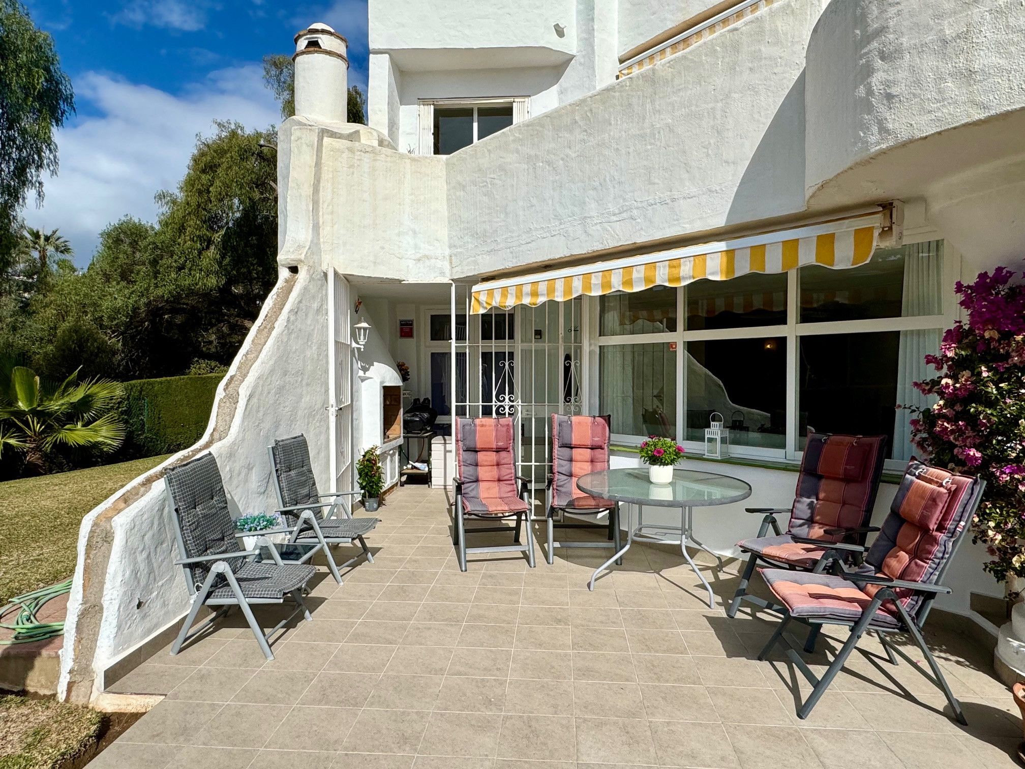 Un patio con mobiliario exterior, una mesa de vidrio y un toldo a rayas amarillas.