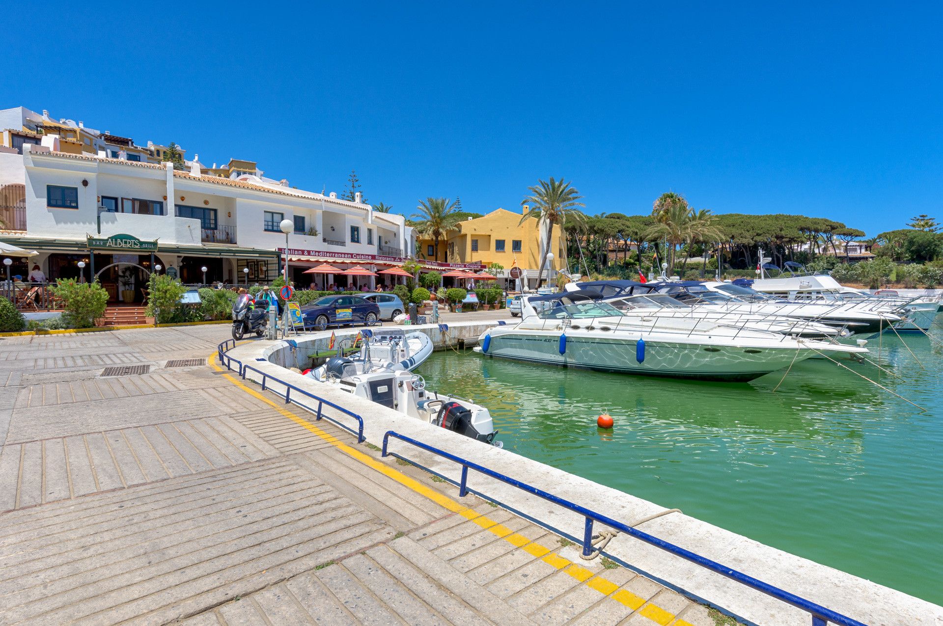 Een jachthaven met verschillende boten aanmeren, een verharde promenade en een restaurant genaamd 'Albert's Grill' met terras.