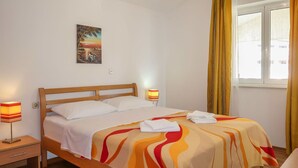 Une chambre d’hôtel avec un lit en bois, du linge de lit blanc, une couverture colorée, une table de chevet avec une lampe, un tableau au mur et une fenêtre avec des rideaux.