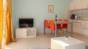 Un salon avec un téléviseur, une table à manger avec des chaises, un coin cuisine avec des placards et une petite plante sur une table.