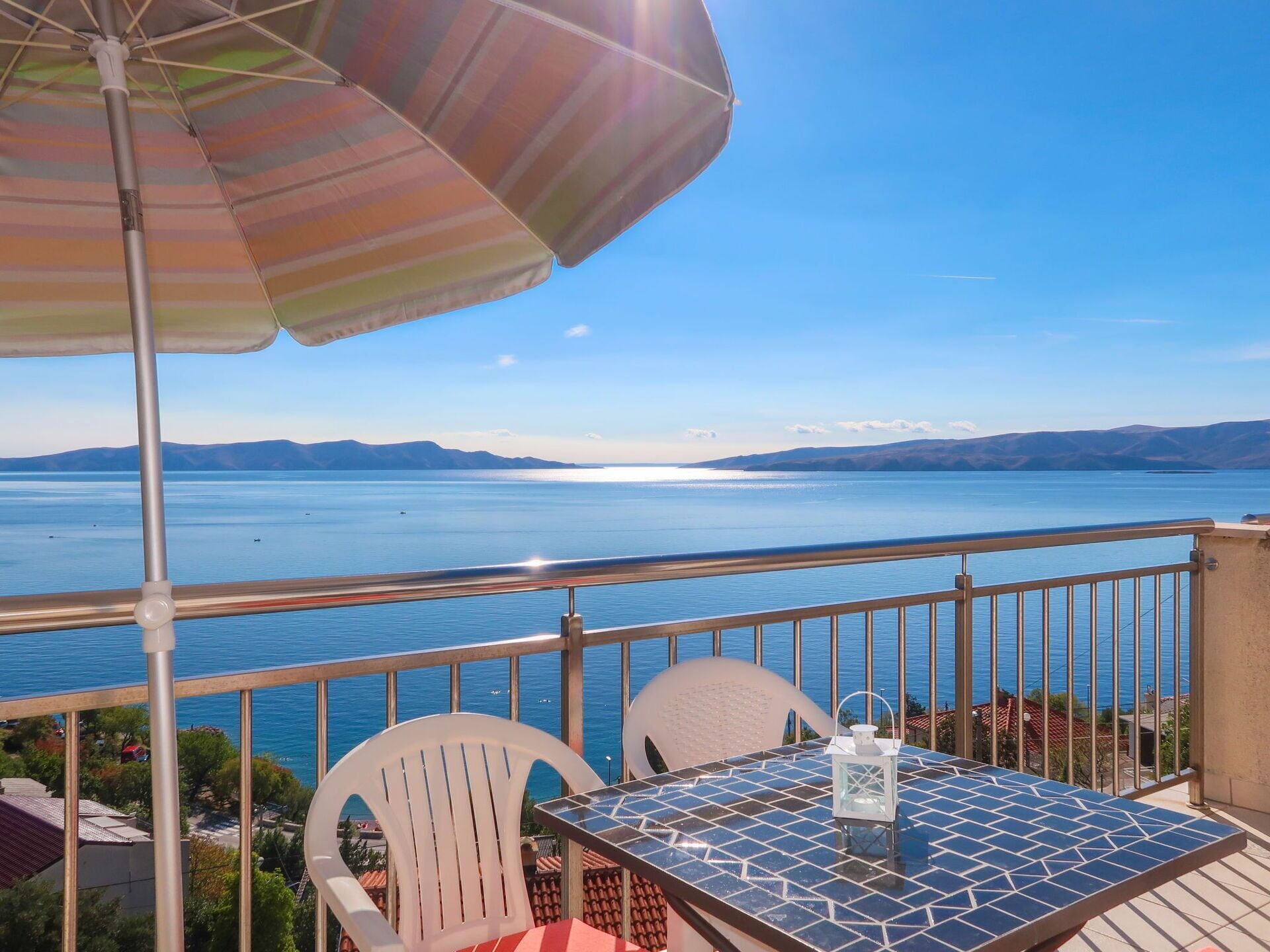A balcony with a table, chairs, and a striped umbrella overlooking a large body of water with distant mountains.