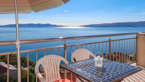 A balcony with a table, chairs, and a striped umbrella overlooking a large body of water with distant mountains. - Lovely apartment for 4 people with WIFI, A/C, TV and terrace (Senj)