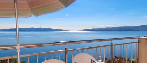 A balcony with a table, chairs, and a striped umbrella overlooking a large body of water with distant mountains.