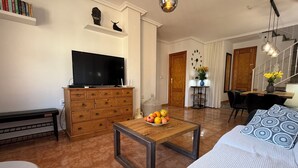 A living room with a sofa, a coffee table with fruit, a TV, a wooden cabinet, and a dining area with chairs and a table. - Familienhaus in Cabo Roig in Strandnähe mit Gemeinschaftspool (VC)