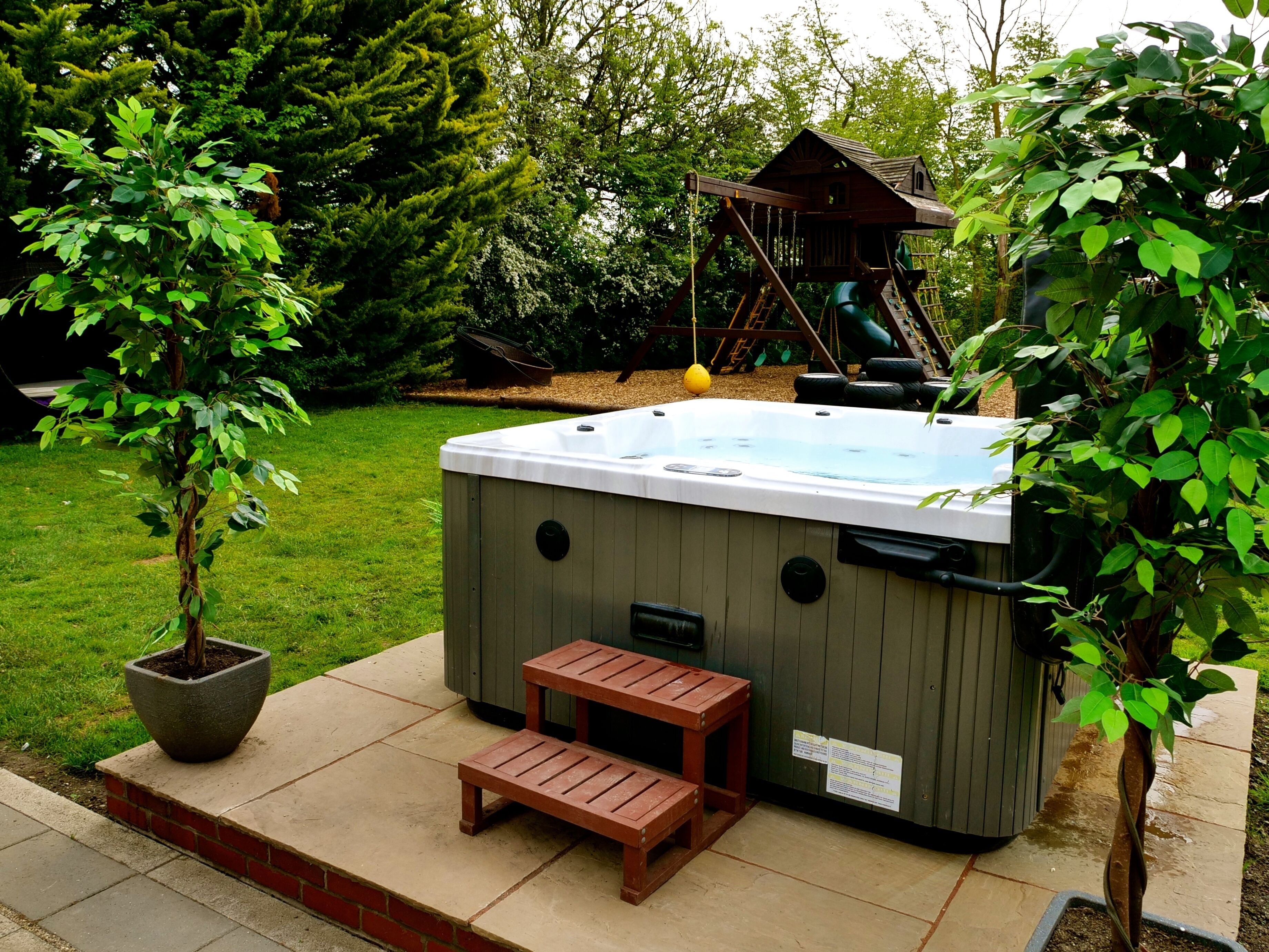 Un jacuzzi con cubierta de madera y un pequeño banco, rodeado de exuberante follaje y una estructura de juego de madera al fondo.