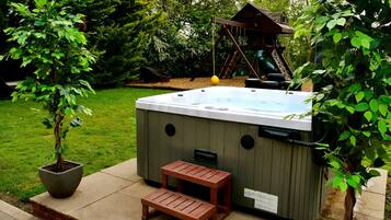 Un jacuzzi con cubierta de madera y un pequeño banco, rodeado de exuberante follaje y una estructura de juego de madera al fondo.