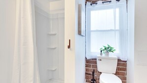 Un baño con ducha blanca, inodoro y pared de ladrillo, y ventana con planta en el alféizar.