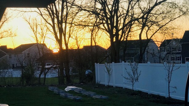 Patio trasero al atardecer con cerca blanca, árboles y césped.