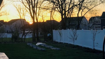 A backyard at sunset with a white fence, trees, and a lawn.