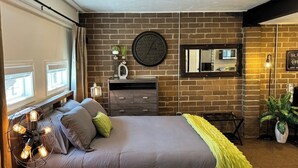 Un dormitorio con pared de ladrillo, un reloj redondo, una cama con almohadas, una mesita de noche, un aparador, un espejo y una planta.