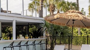 Uma área à beira da piscina com bar, espreguiçadeiras e um guarda-sol de palha.