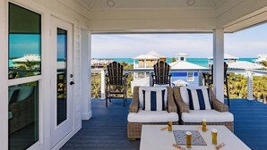 A covered outdoor seating area with wicker furniture and a table, overlooking a beach and ocean. - Lookout Escape: Heated Pool, Golf Cart, Outdoor Bar, Gulf View (North Captiva)