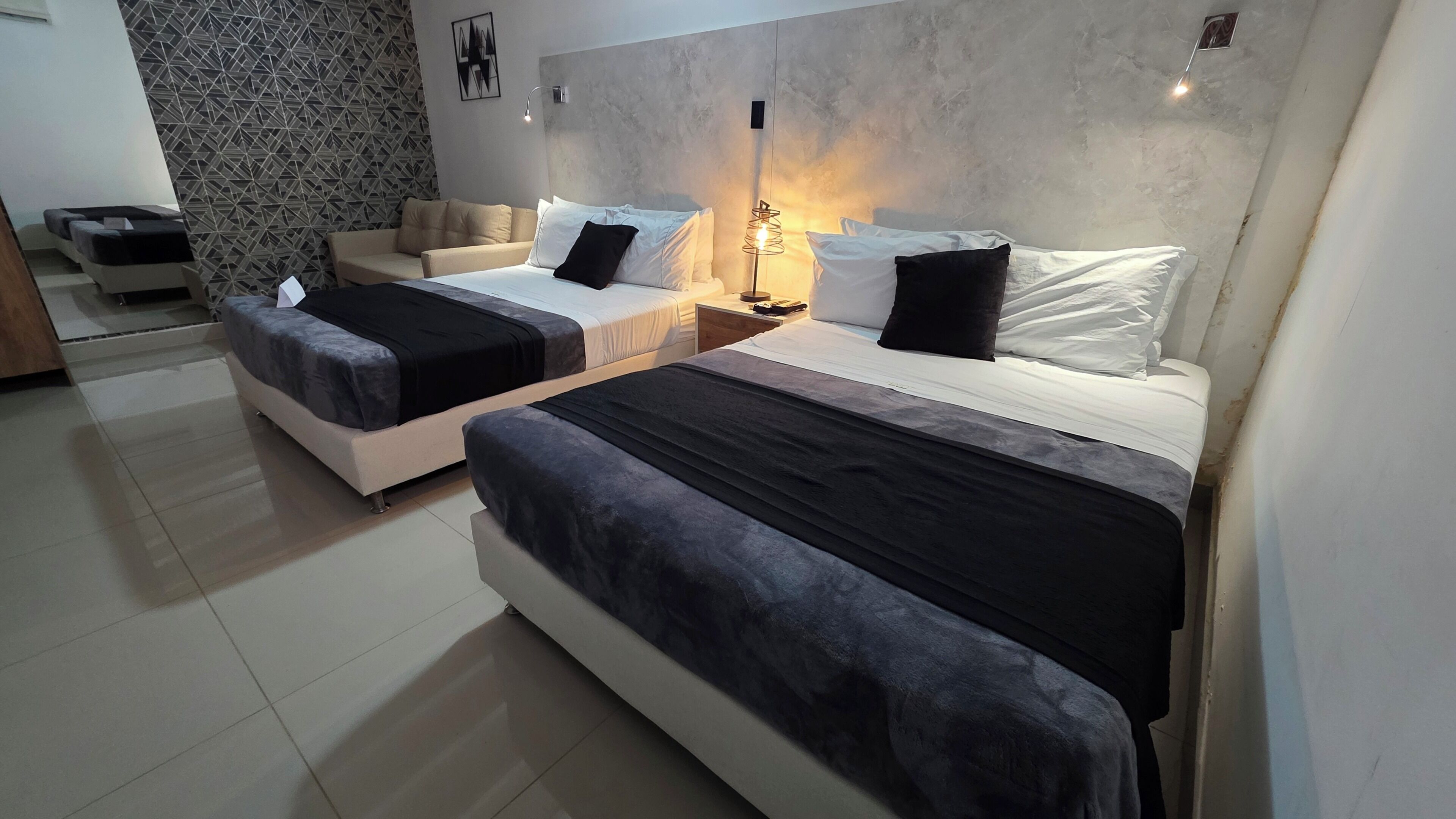 Quarto de hotel com duas camas, um sofá, parede estampada e luminária moderna.