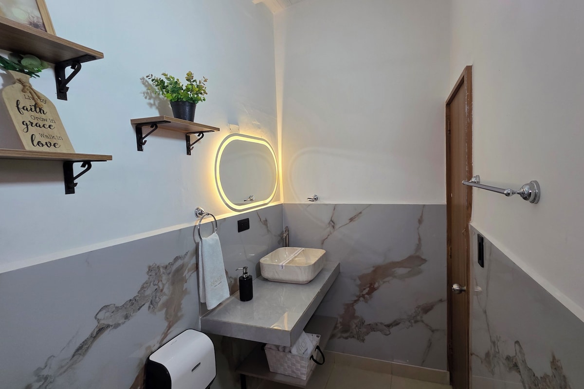 Un bany amb una paret de marbre, un lavabo amb mirall, un suport de tovalloles, una planta i una estant amb un cartell.