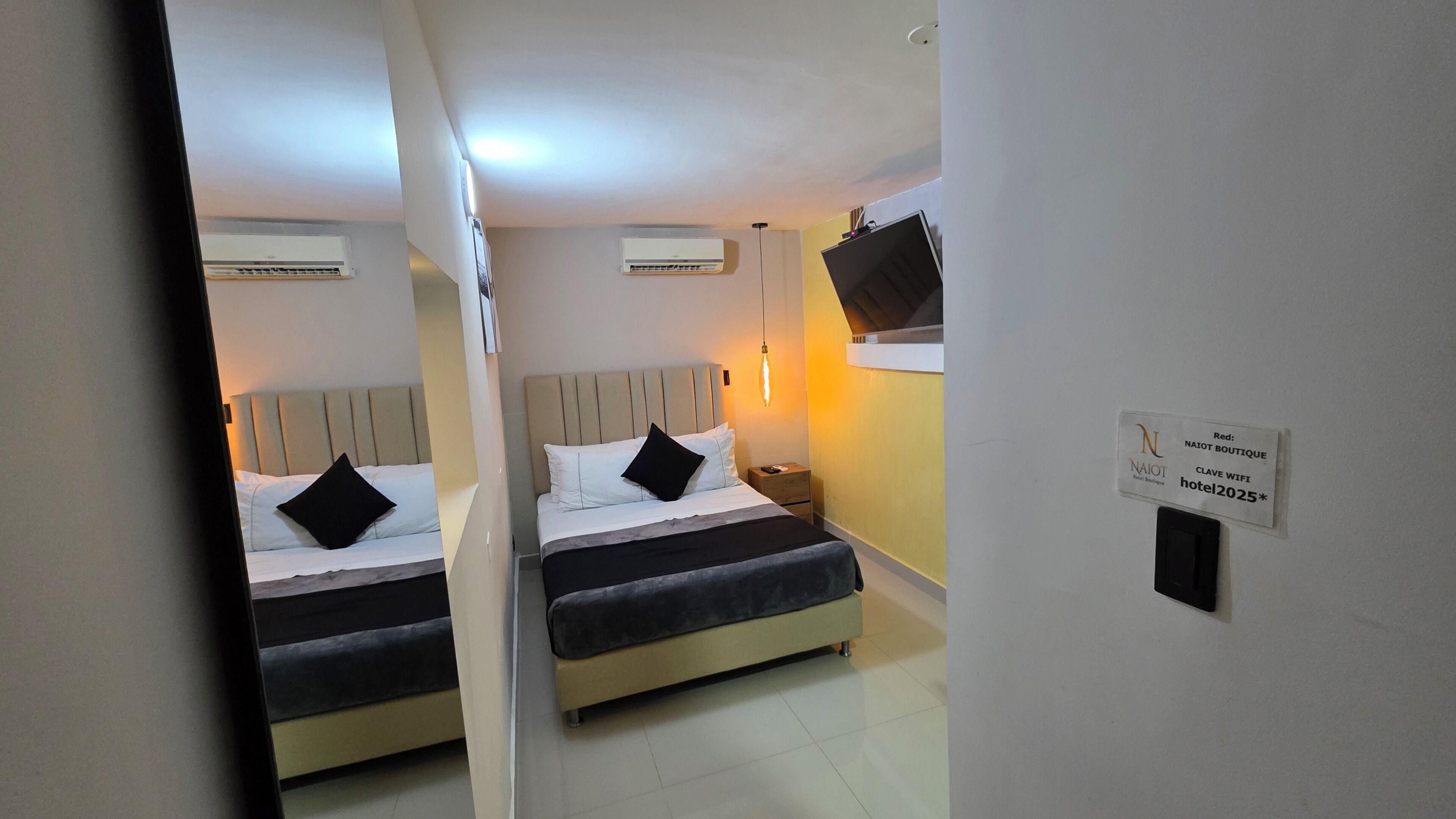 Quarto de hotel com duas camas, uma mesa de cabeceira, um aparelho de ar condicionado e uma televisão fixada na parede.