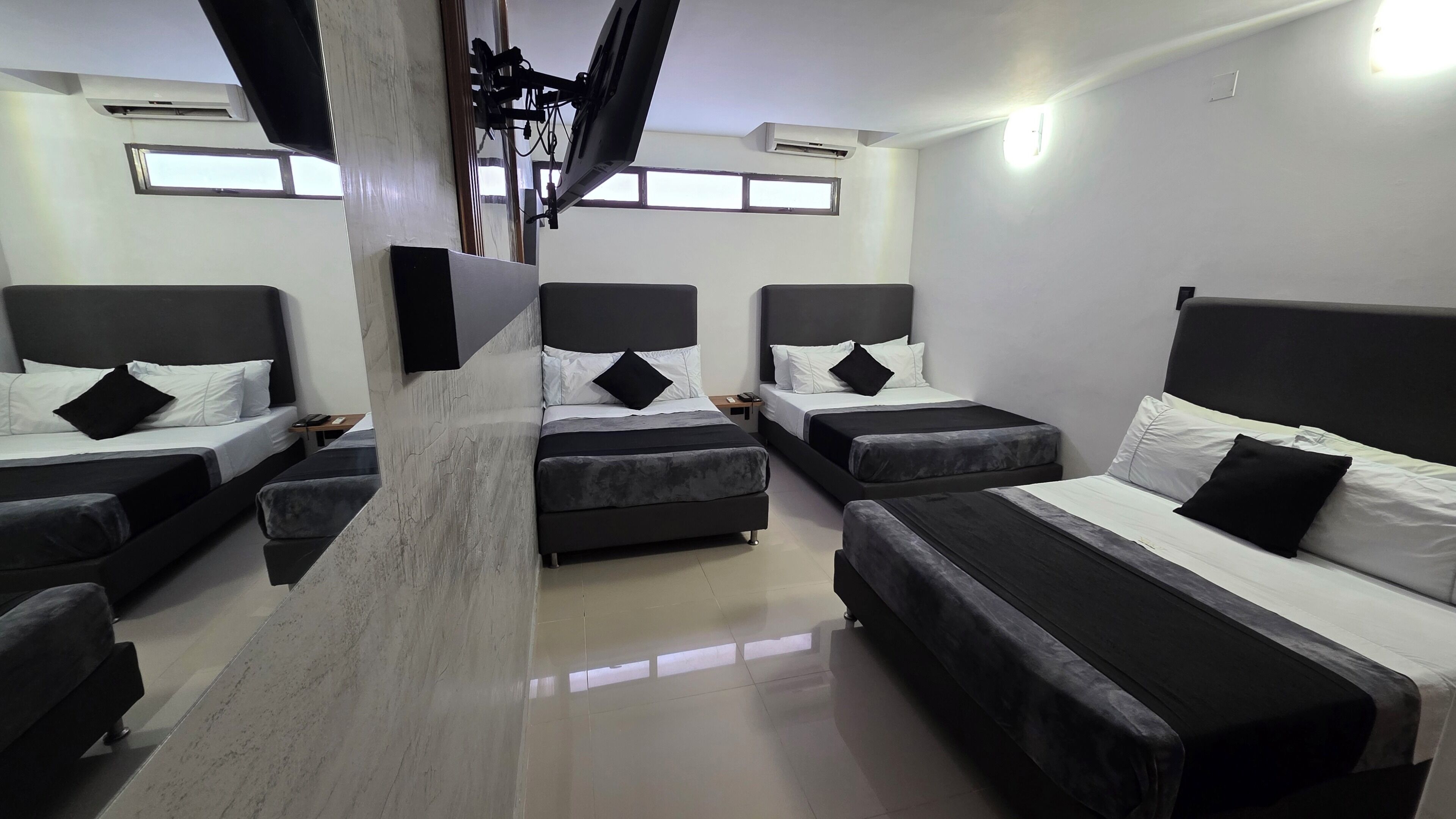 Quarto de hotel com quatro camas dispostas em formato de U, uma TV de tela plana na parede e um ventilador de teto.