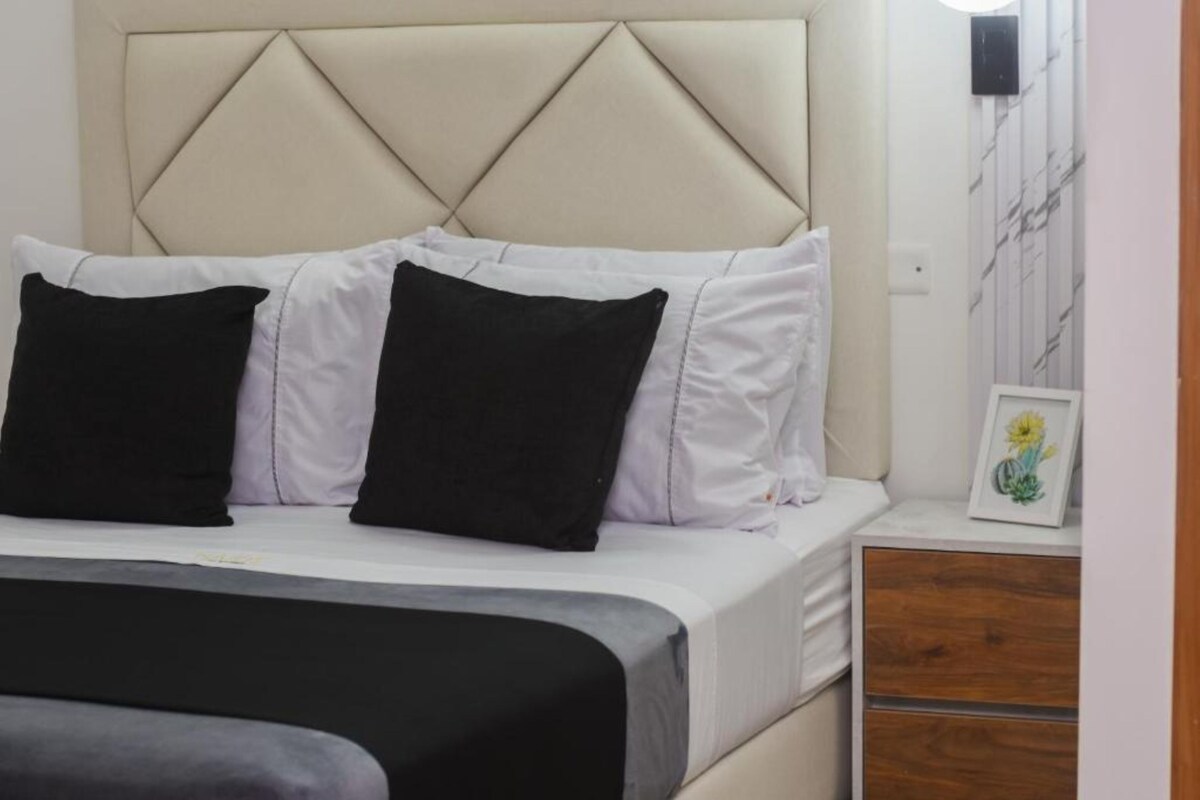 A bed with a headboard, white and black pillows, and a wooden nightstand with a framed picture.