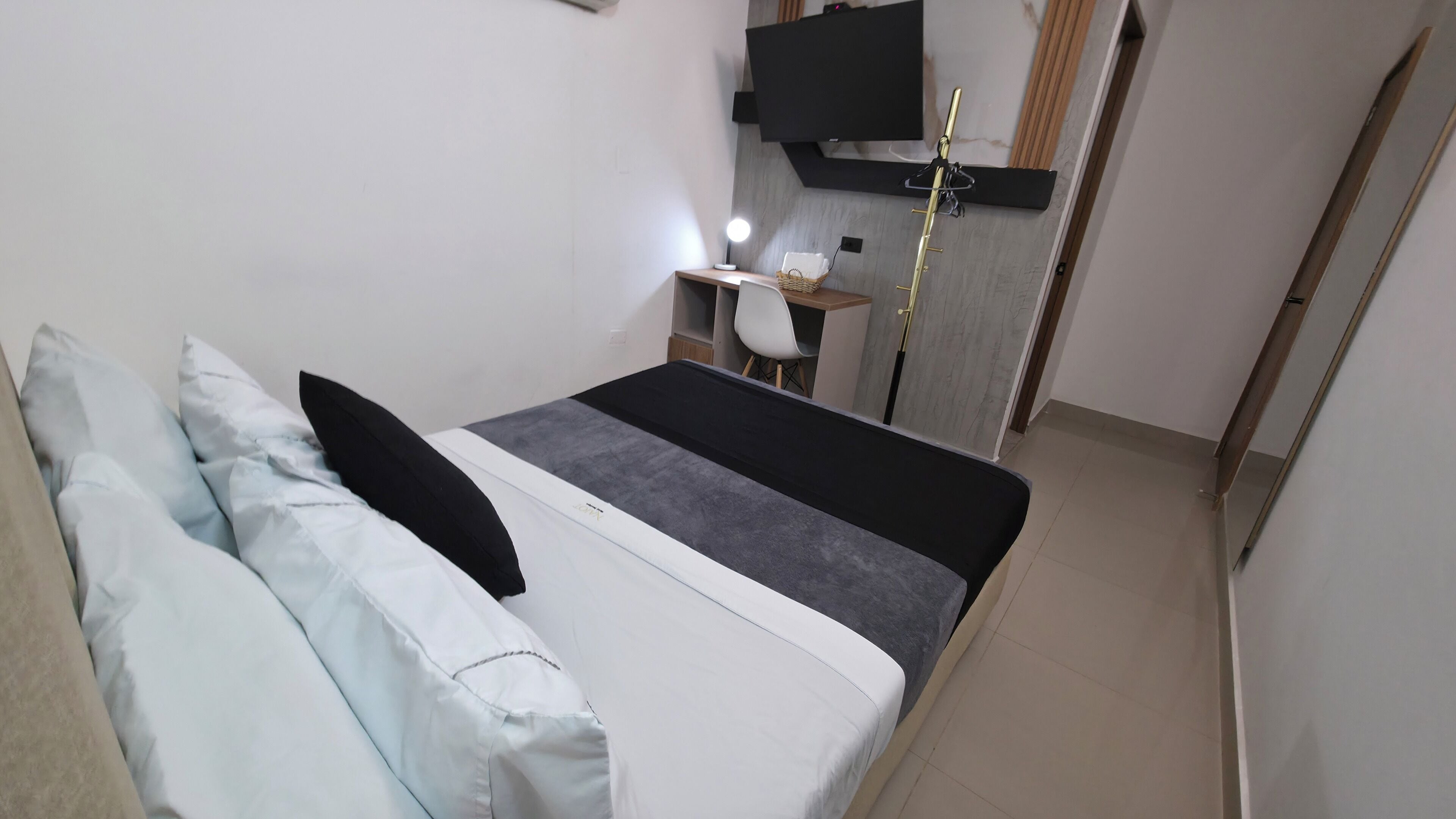 Quarto com cama, almofadas, escrivaninha com cadeira, abajur e televisão na parede.