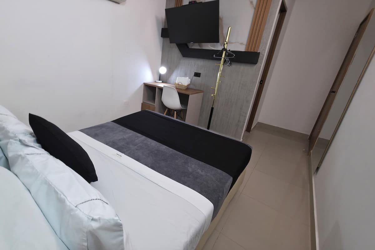 A bedroom with a bed, pillows, a desk with a chair, a lamp, and a television mounted on the wall.