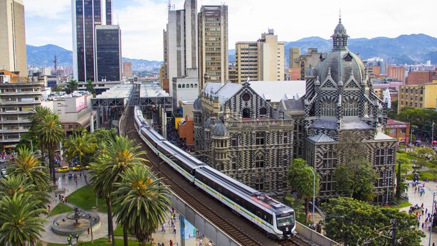 Una estació de tren moderna amb un tren d'alta velocitat, envoltada d'edificis urbans i palmeres.
