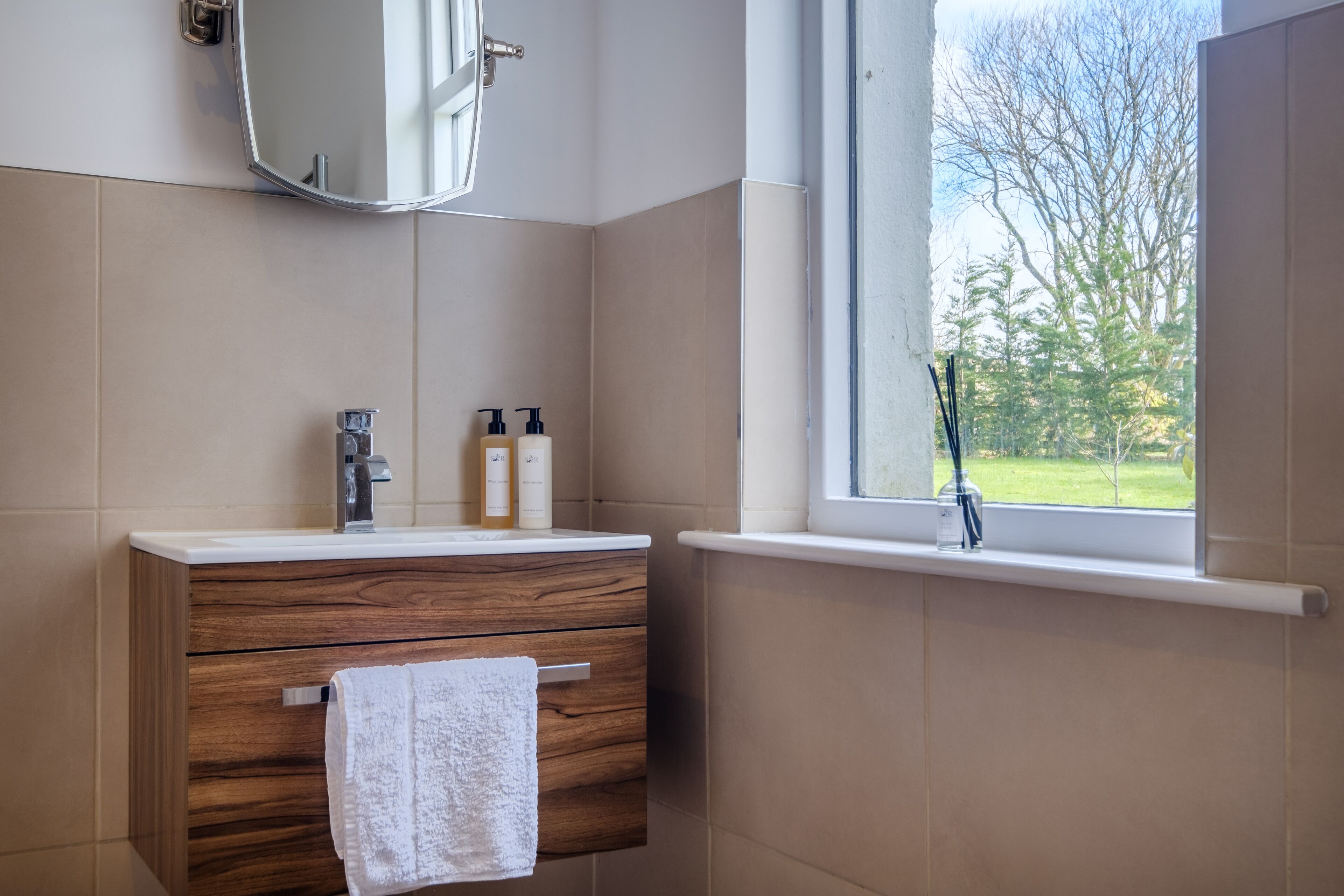 Een badkamer met een houten wastafelmeubel, een spiegel, een raam met uitzicht op bomen en toiletartikelen op het aanrecht.