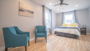 Une chambre à coucher avec un lit, deux chaises, un ventilateur de plafond et une fenêtre avec des rideaux.