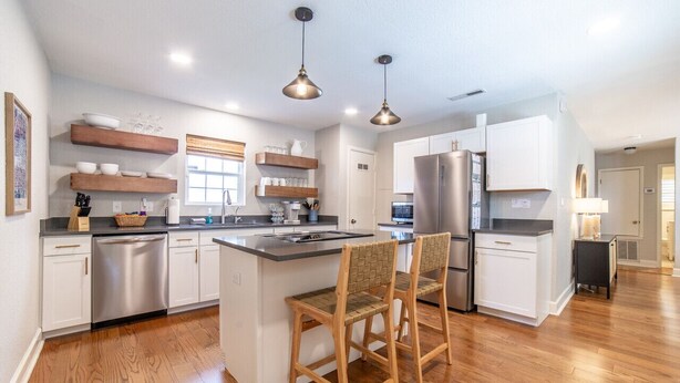 A modern kitchen with white cabinets, a stainless steel dishwasher, a large island with bar stools, and pendant lighting. - The Garland Getaway - Short Walk to UofA (Fayetteville)
