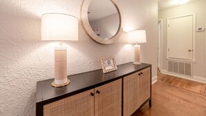 A hallway with a textured wall, a round mirror, two wall-mounted lamps, a framed picture, and a dark wooden console with wicker doors. - The Garland Getaway - Short Walk to UofA (Fayetteville)
