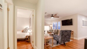A room with hardwood flooring, a bed, a gray armchair, a small table with a lamp, and a ceiling fan. - The Garland Getaway - Short Walk to UofA (Fayetteville)