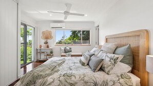 A bedroom with a large bed, a ceiling fan, a window with a view of greenery, and a bedside table with decorative items. - TreasureView Beach House #9 -Entire property (coolum)