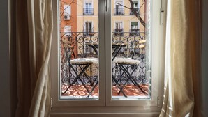 Vista per una finestra amb barana de balcó metàl·lic, una cadira i un edifici amb diversos balcons al fons.
