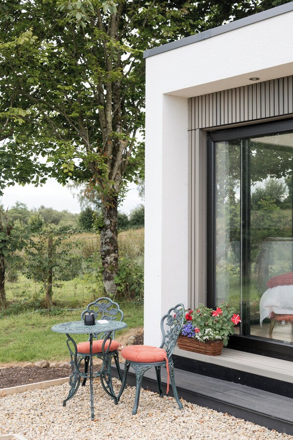 A modern outdoor seating area with a table and two chairs, a wicker basket with flowers, and a large window.