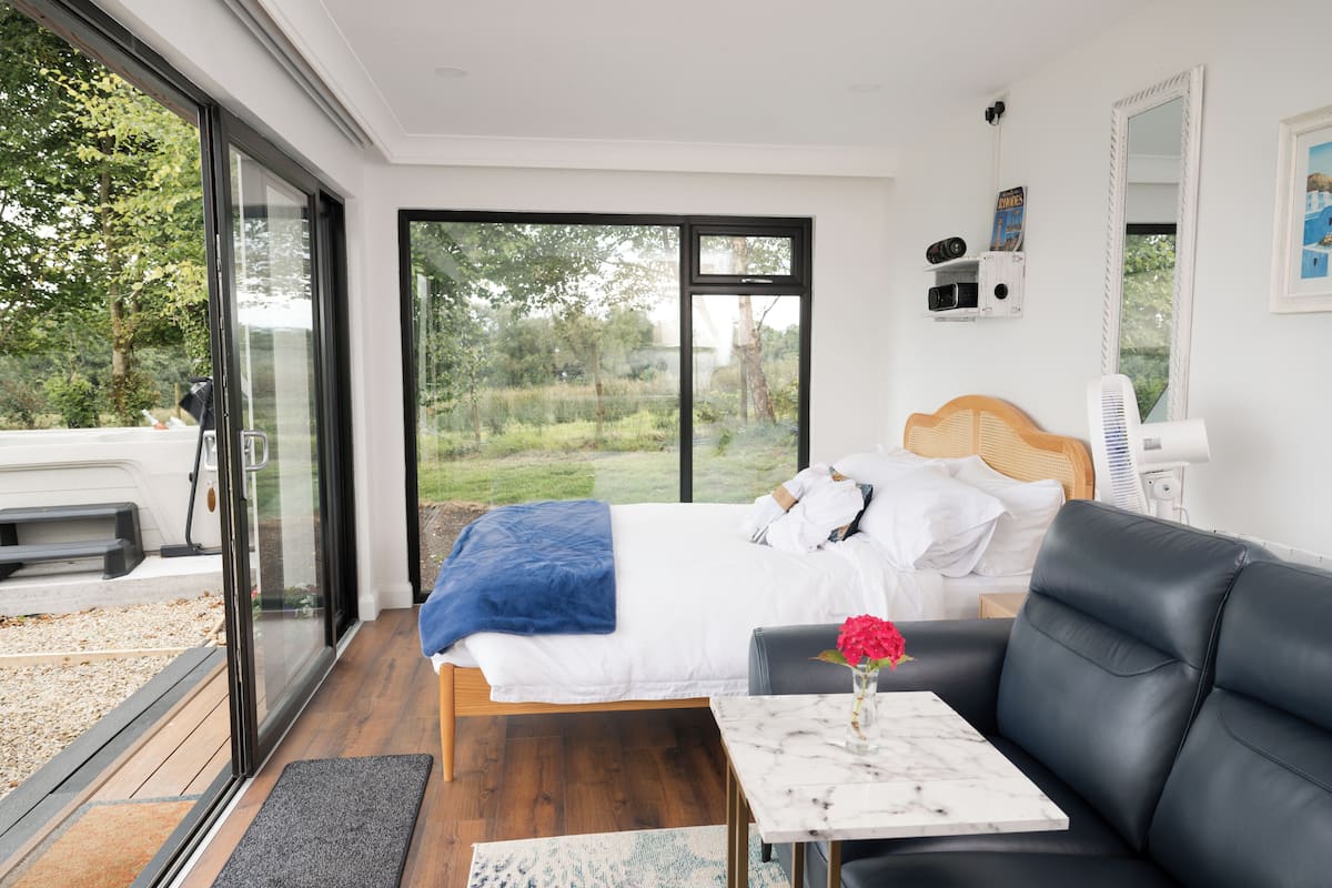 Ein modernes Schlafzimmer mit einem großen Bett, einer schwarzen Ledersofa, einem kleinen Tisch mit einer Vase und Blick ins Freie durch große Fenster.