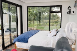 Une chambre avec une grande porte-fenêtre coulissante donnant sur un jardin, un lit avec une literie blanche, un plaid bleu et un ventilateur blanc.