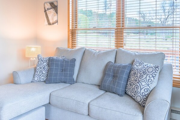 A light grey sofa with patterned cushions, a floor lamp, and a window with blinds.