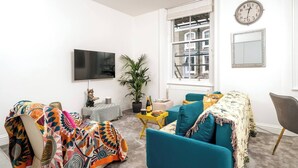 Ein modernes Wohnzimmer mit einem Flachbildfernseher, einer großen Topfpflanze, Möbeln in Türkis und Gelb sowie Blick auf die Stadt durch das Fenster.