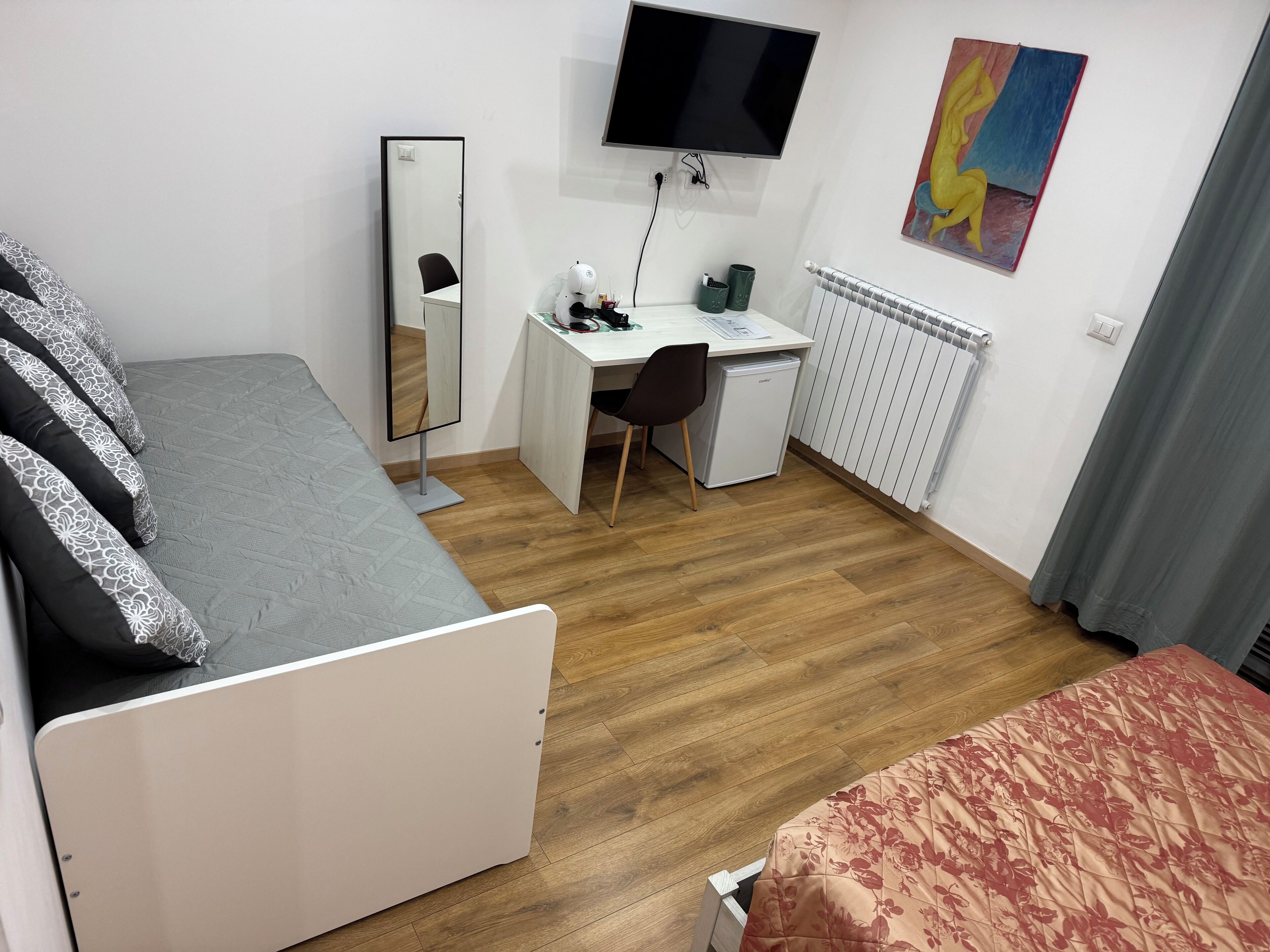 A bedroom with a bed, a desk, a chair, a TV, a radiator, and a painting on the wall.