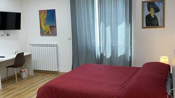 A bedroom with a bed, a desk, a chair, a TV, and artwork on the walls.