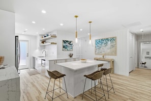 Une cuisine moderne avec des meubles blancs, un îlot central doté de tabourets, un éclairage suspendu et des œuvres d’art accrochées aux murs.