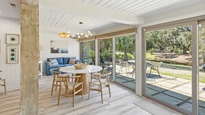 Un comedor amplio con una mesa redonda y sillas, un sofá azul y un gran ventanal corredera que da a un patio exterior con piscina y jardín.