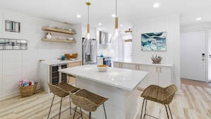 Una cocina moderna con armarios blancos, una isla central, taburetes de bar, luces colgantes y un cuadro en la pared.