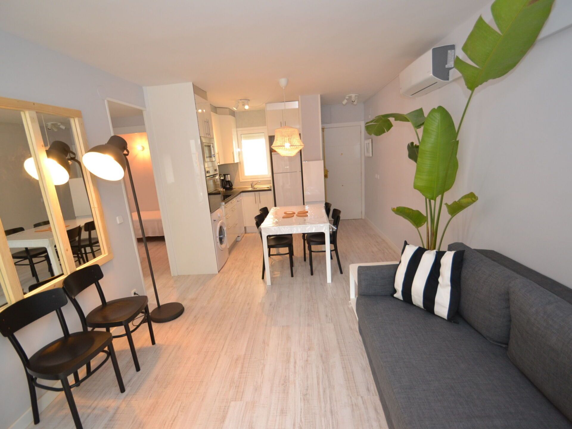 Ein modernes Wohnzimmer mit grauem Sofa, einem Esstisch mit Stühlen, einem großen Spiegel und einer Küche im Hintergrund.