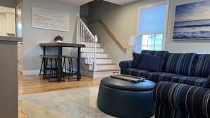 A living room with a striped sofa, a round ottoman, a coffee table, a bar stool, a staircase, and a painting of the ocean. - Unwind Oceanside
Private single family home (Hampton)