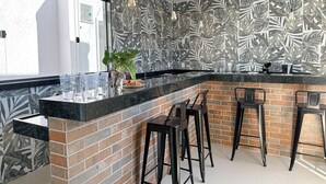 A kitchen with a black marble countertop, a brick backsplash, and a large window. There are three bar stools and a potted plant on the counter. - CoVívere - RJ (Rio de Janeiro)