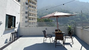A balcony with a table, chairs, and an umbrella, overlooking a mountain and a building. - CoVívere - RJ (Rio de Janeiro)
