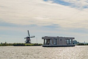 A modern building with a flat roof and large windows, situated on a body of water with a windmill in the background.