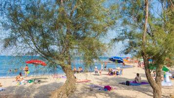 Una playa con gente tomando el sol y nadando, árboles y sombrillas.