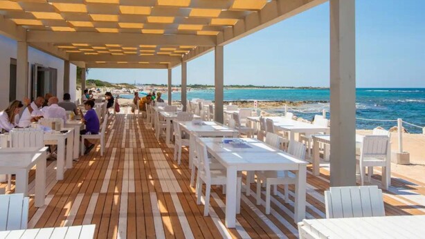 Un área de comedor al aire libre con mesas y sillas blancas, una terraza de madera y vistas a la playa y al océano.